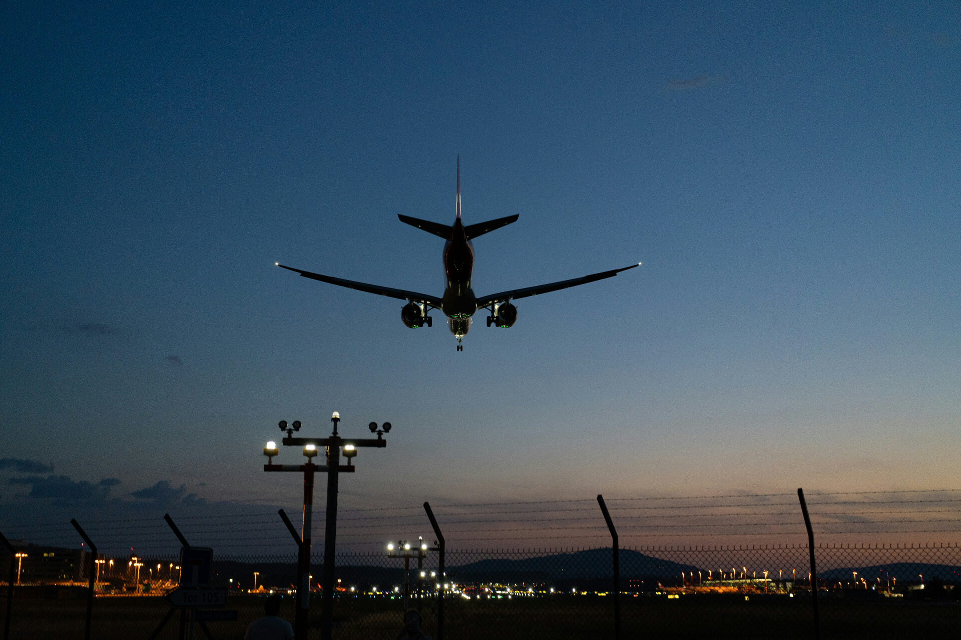 A wide-body airliner on final approach at dusk over Zurich Airport, with runway lights glowing along the perimeter.