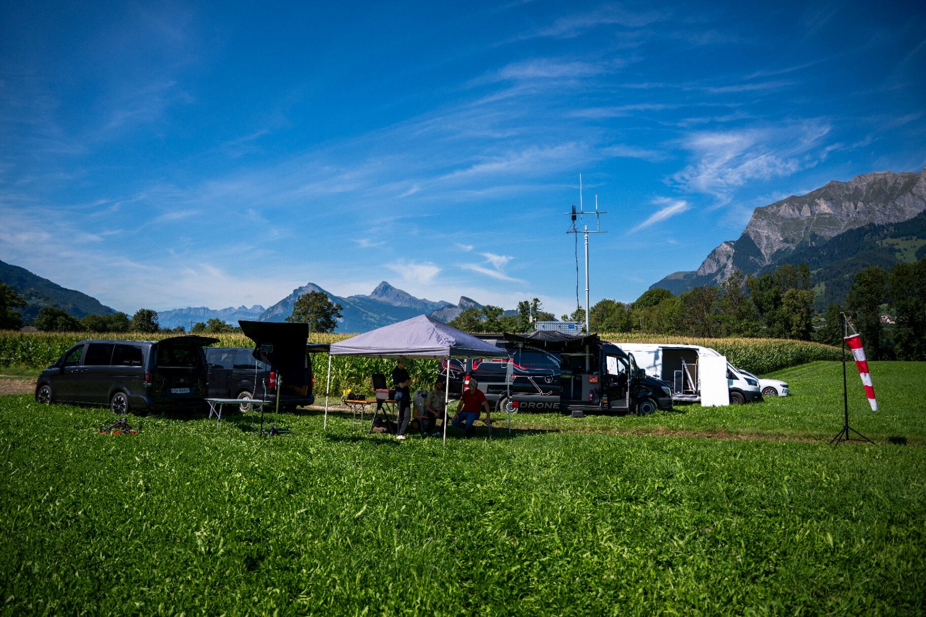 Drone base camp with vans, antenna mast and tents in an alpine field below the Swiss Alps