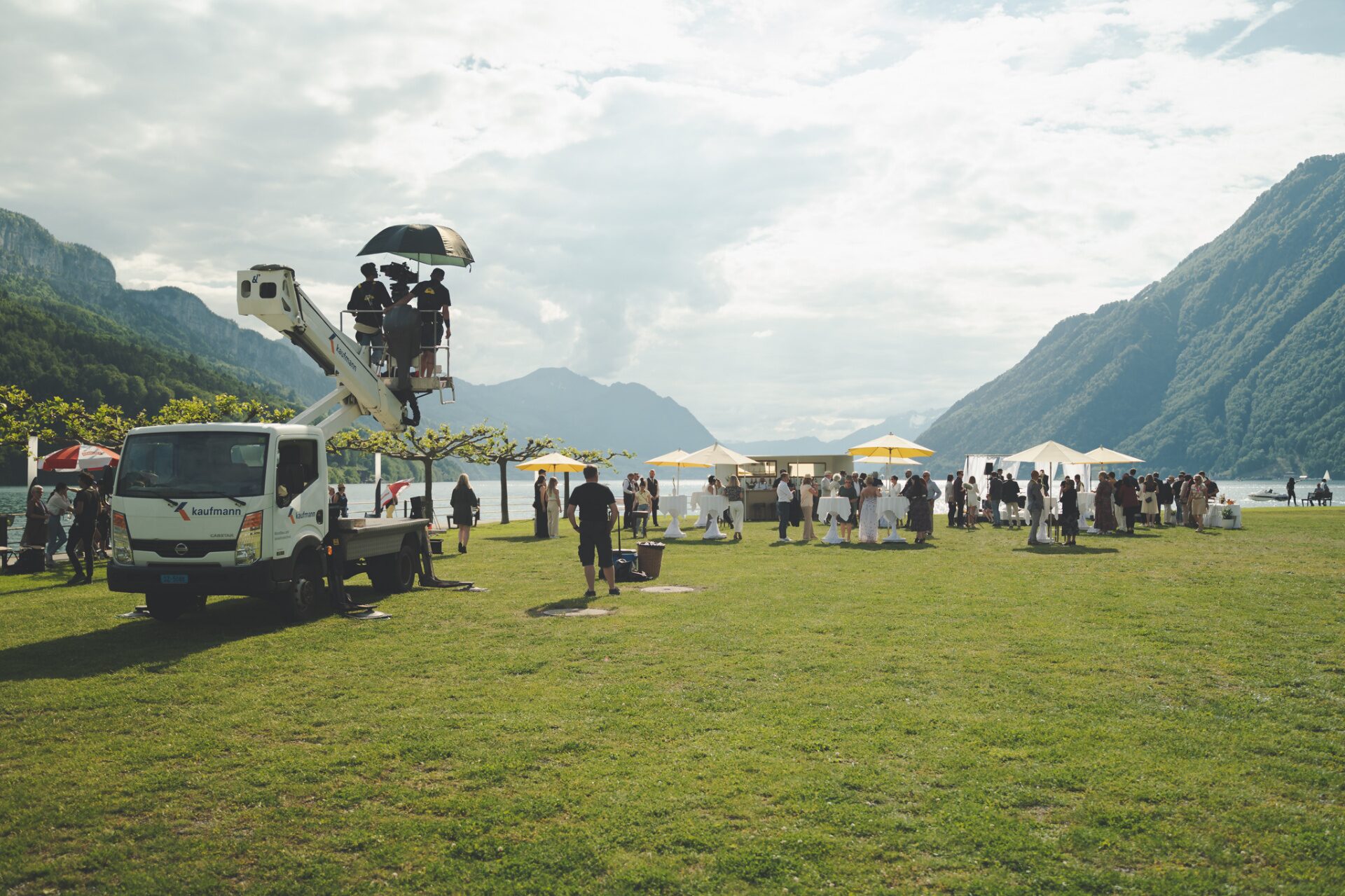 Camera crew on a cherry-picker lift filming a lakeside event between Swiss mountain ridges
